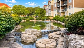 A landscaped garden with a pond, rock formations, and a multi-story building in the background.