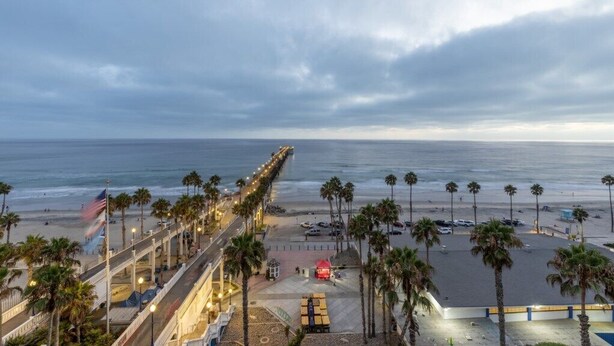 A beachside promenade with a long pier extending into the ocean, palm trees lining the path, and a parking area with cars.