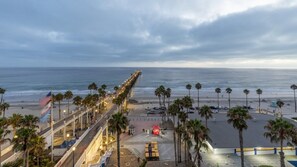 A beachside promenade with a long pier extending into the ocean, palm trees lining the path, and a parking area with cars.