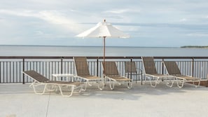 Relax on the seaside terrace with a row of sun loungers and a shaded area under a single umbrella, taking in the views of the calm body of water and distant shoreline.