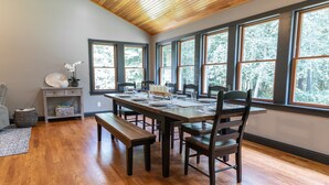 A dining area with a wooden table, benches, and chairs. There are large windows with a view of trees. A small shelf with decorative items is visible. - Perfect getaway for large families who love the woods and beach!! (Woodbine)
