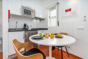 Een compact keuken met een magnetron, waterkoker en broodrooster. Er is een eettafel met borden, bestek en een fruitschaal. Een rood noodbordje hangt aan de muur.