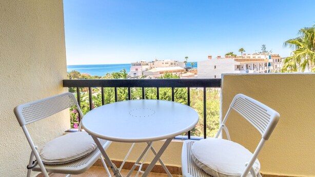 A balcony with two white chairs and a small round table, overlooking a coastal view with buildings and greenery. - #063 Beach Flat by Home Holidays (Albufeira)