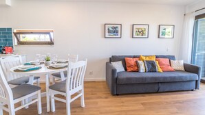 A modern living room with a grey sofa, white chairs, a dining table set for four, and a kitchen area with a window. - #063 Beach Flat by Home Holidays (Albufeira)