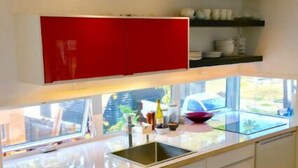 Eine moderne Küche mit weißen Schränken, einem roten Oberschrank, einer Glas Arbeitsplatte und einem Fenster mit Blick ins Freie.
