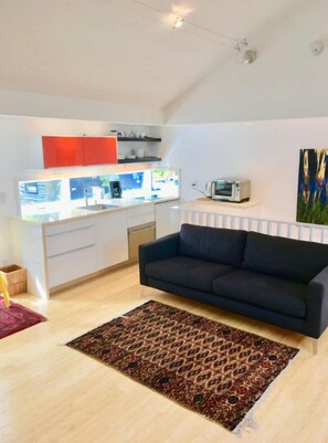 A modern living room with a dark sofa, a patterned rug, and a kitchen area with white cabinets and a red accent wall. - Bright Madison Valley Guest House (Seattle)