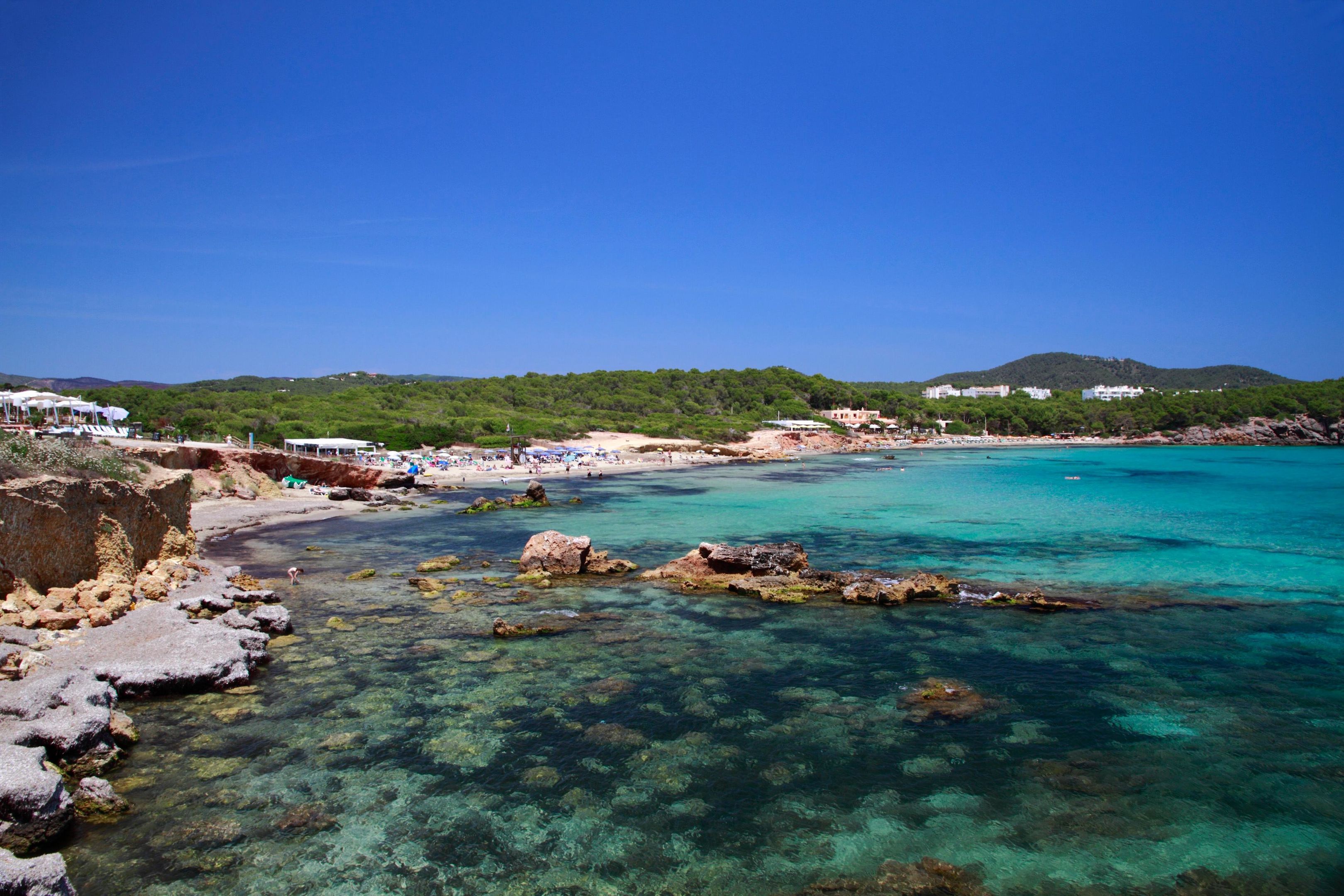 A coastal beach with clear water, rocky shore, and a distant hill.