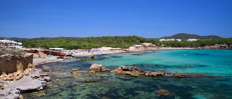 A coastal beach with clear water, rocky shore, and a distant hill.