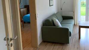 A room with a green sofa, a wooden floor, a door leading to another room, and a window with curtains.