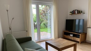 A living room with a green sofa, a wooden coffee table, a TV on a wooden stand, and a sliding glass door leading to a garden.
