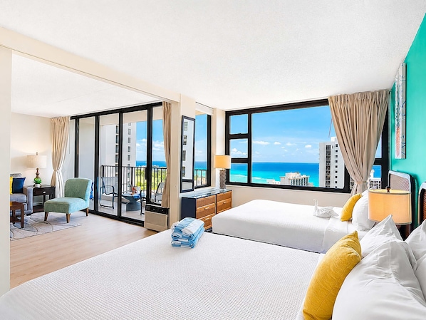 A hotel room with two beds, a balcony, a desk, and a view of the ocean.