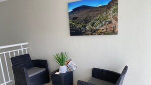 En balkon med to fletstole, et lille bord med en plante og tidsskrifter, samt et indrammet billede af et bjerglandskab.