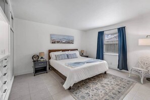 Une chambre à coucher avec un lit, des tables de chevet, une lampe, une chaise, une fenêtre avec des rideaux et un tableau au mur.
