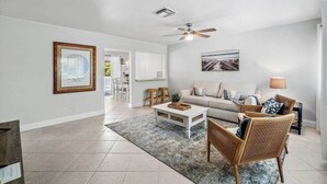 Un vaste salon avec un canapé blanc, une table basse et des fauteuils en osier. On y trouve un ventilateur de plafond, un tableau encadré au mur et une vue sur la salle à manger.