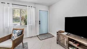 Une pièce avec un téléviseur posé sur un meuble en bois, une chaise en osier, une porte bleue et une fenêtre ornée de rideaux légers.