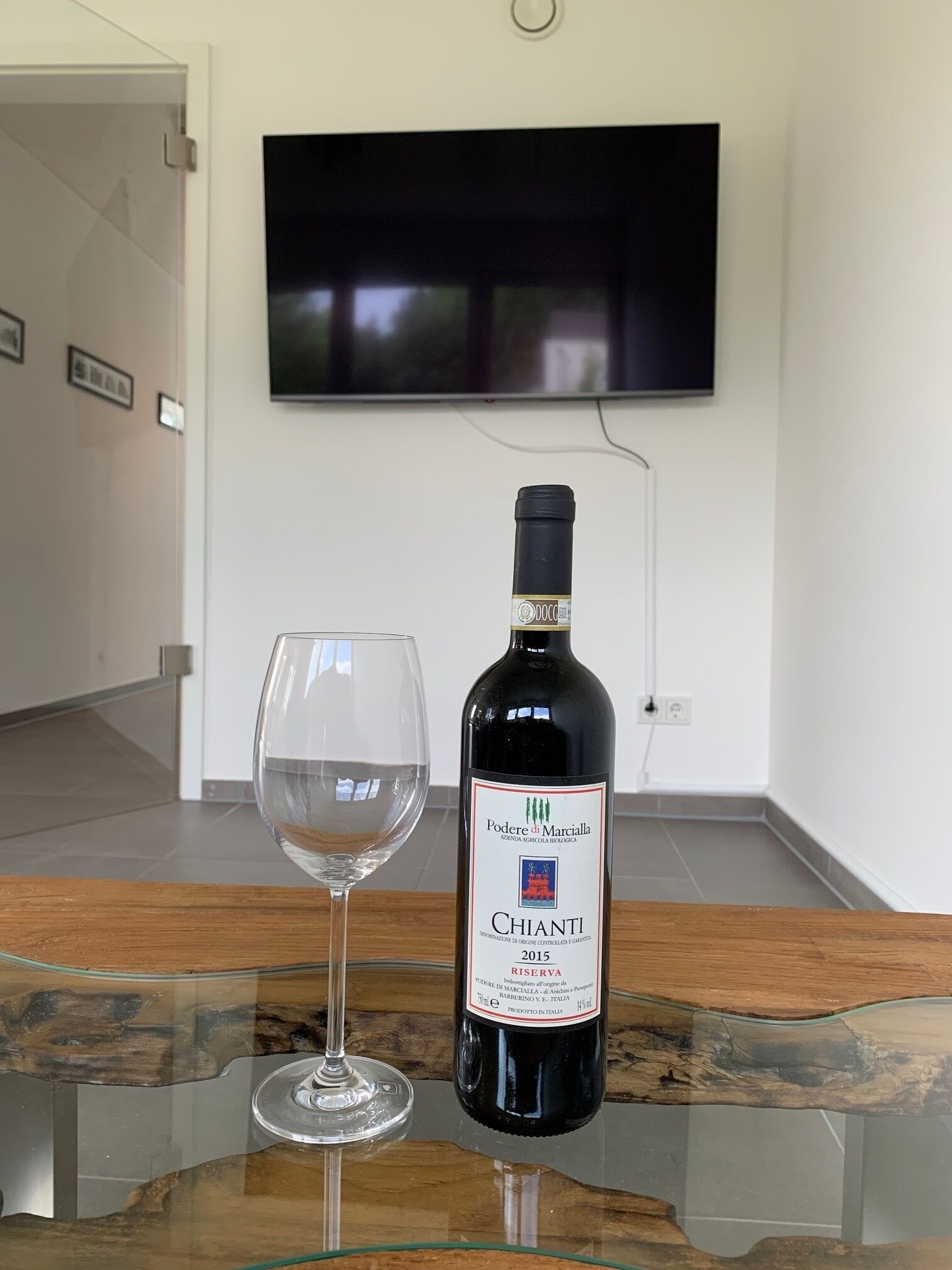 A bottle of Chianti wine and a wine glass on a glass table in a room with a television mounted on the wall.