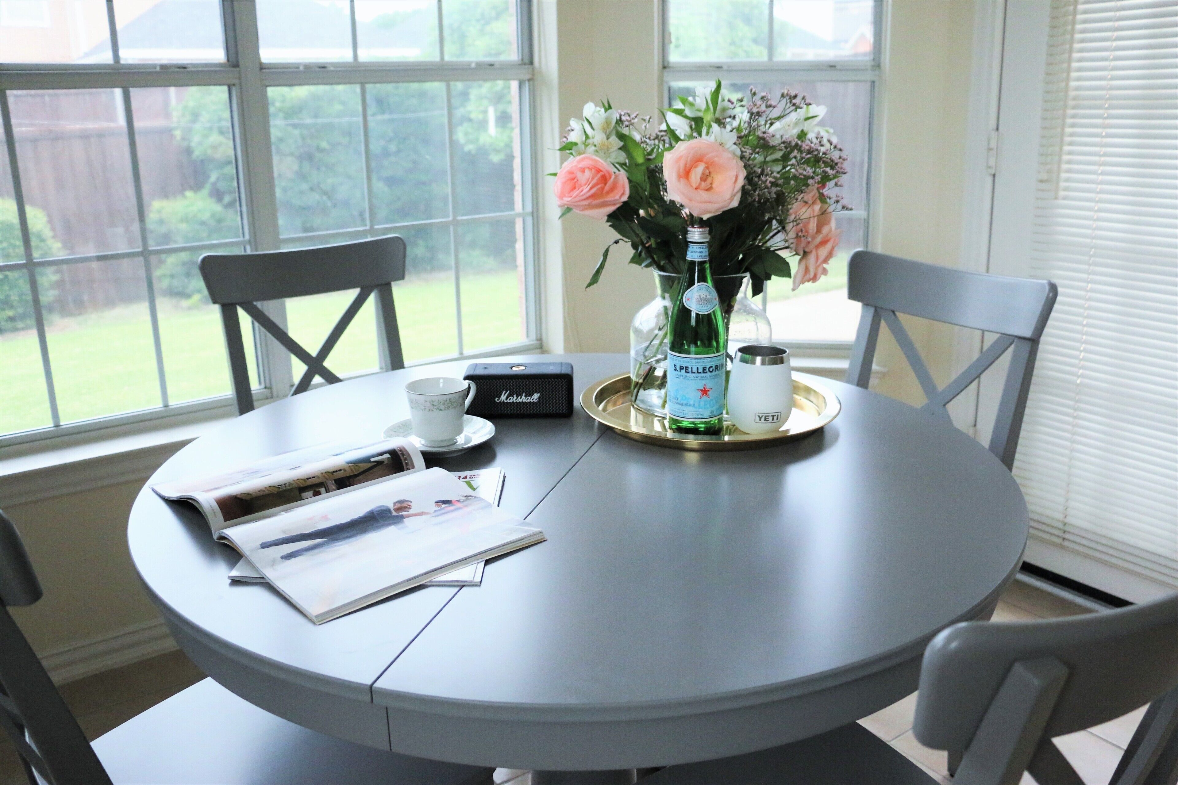 A round dining table with a magazine, a small speaker, a tray with a bottle and a cup, and a vase with flowers.