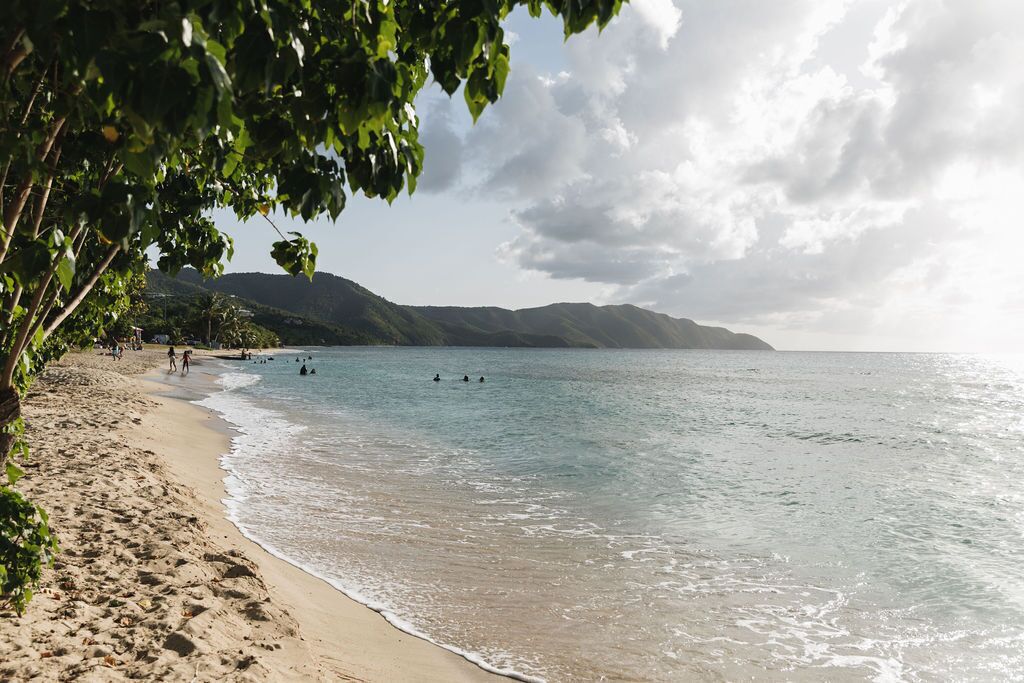 A beach with clear water, people swimming, and mountains in the background.