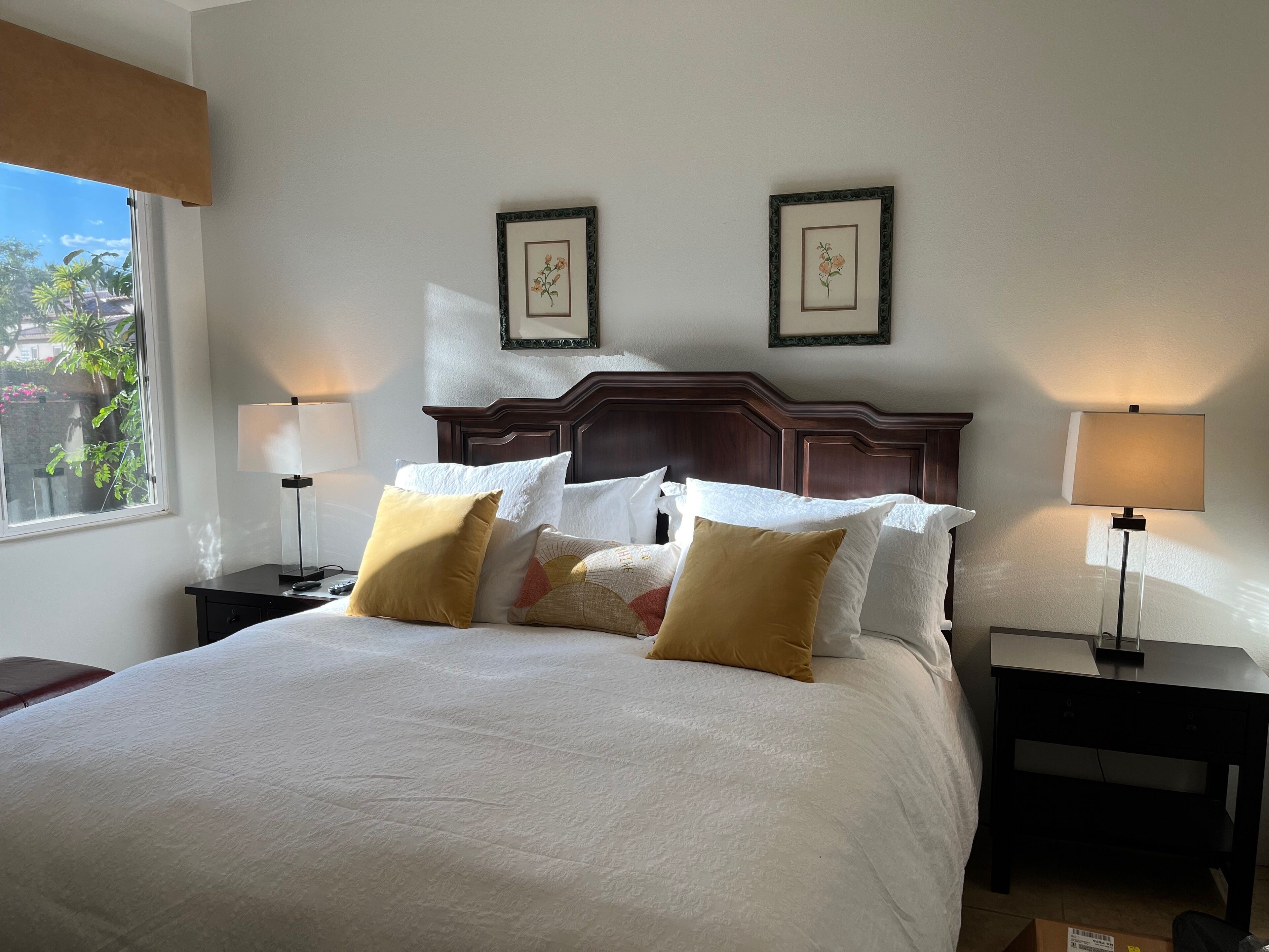A neatly made bed with white and mustard pillows, two framed floral pictures on the wall, and two bedside lamps.