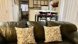 Een donkerbruine leren bank met twee gestreepte kussens, een keuken met witte kasten en een eettafel met stoelen op de achtergrond.