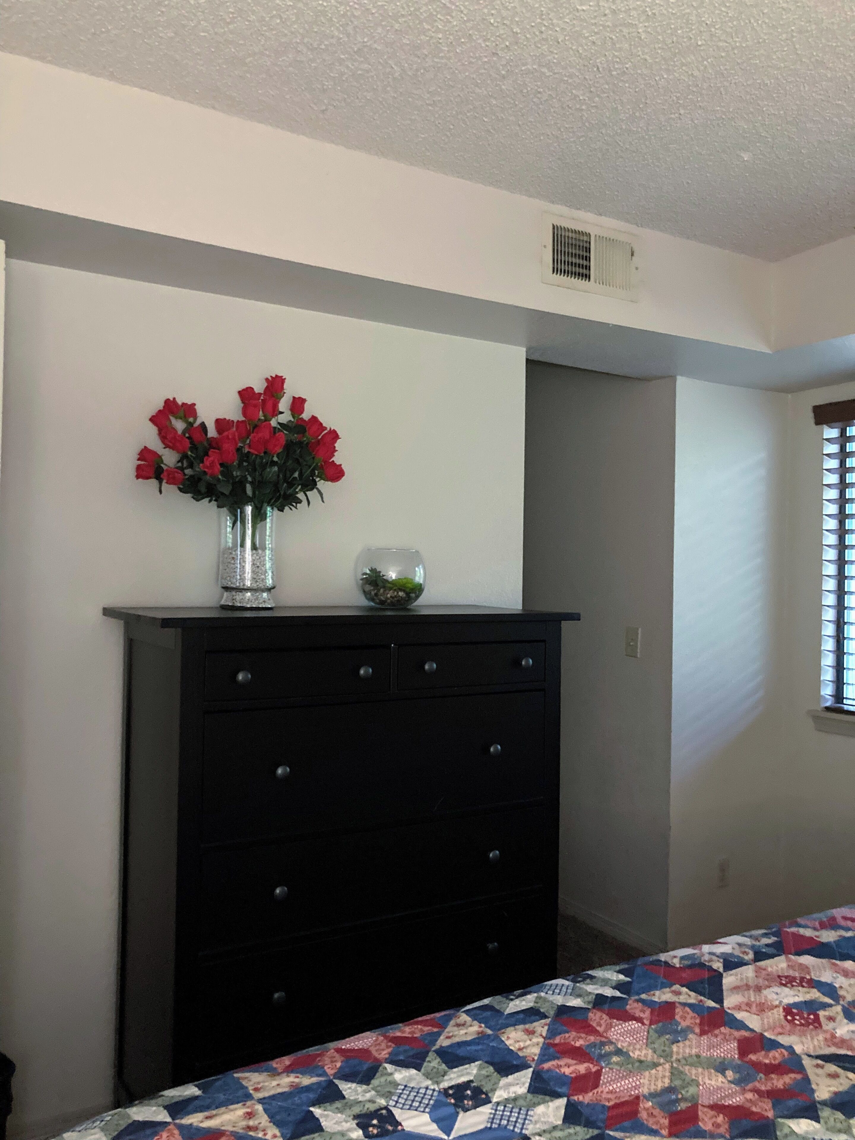 A bedroom with a bed, a dresser, a vase with red flowers, and a window with blinds.