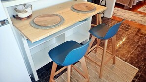 Et lille køkkenområde med en træbordplade, to blå stole og en vævet måtte.
