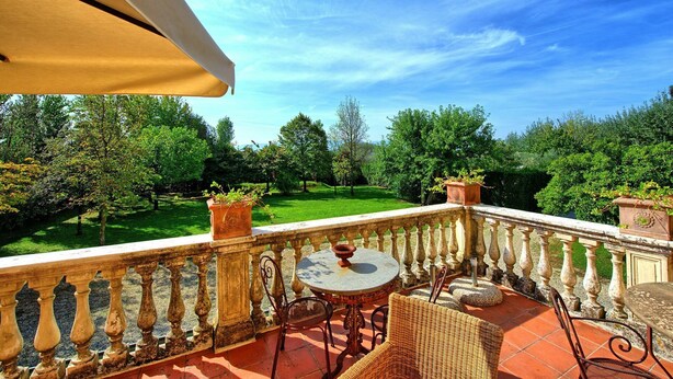 Ein Balkon mit rundem Tisch und Korbstühlen, mit Blick auf einen gepflegten Garten mit Bäumen und blauem Himmel.