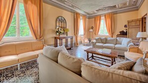 Ein geräumiges Wohnzimmer mit einer großen Couch, einer kleineren Couch und einem Couchtisch. Zwei Fenster mit Vorhängen, eine dekorative Decke und ein gefliester Boden sind ebenfalls vorhanden.