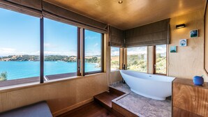 A modern bathroom with a freestanding bathtub, large windows offering a scenic view of a coastal landscape. - Designer living with stunning views across the bay, a spa and coast walks. (Waiheke Island)