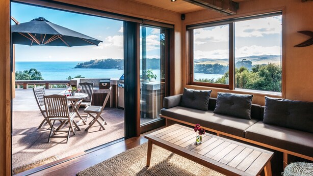 A spacious room with a large sliding glass door leading to a balcony overlooking a scenic view of the sea and hills. Inside, there is a wooden sofa, a coffee table, and a dining area with chairs and a table. - Designer living with stunning views across the bay, a spa and coast walks. (Waiheke Island)