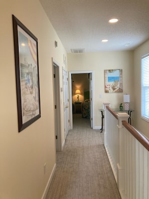 A hallway with a carpeted floor, a framed picture on the left, a lamp in the middle, and a window on the right.