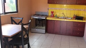A compact kitchen with a gas stove, a sink, and a small countertop. There is a chair and a table in the dining area. - Las Palmas (Ejido Plutarco Elias Calles)