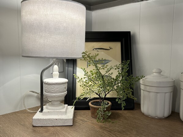 Ein Tisch mit einer Lampe, einem gerahmten Bild, einer Topfpflanze und einem weißen Behälter.