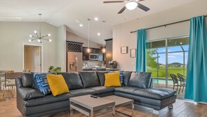 A modern living room with a sectional sofa, a coffee table, and a view of the kitchen and outdoor patio. - Modern Coastal 3 Bedroom Villa with Salt Water Heated Pool & Office! (Cape Coral)