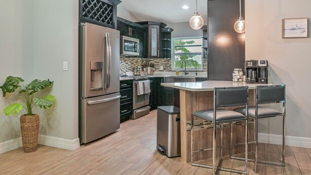 A modern kitchen with stainless steel appliances, a central island with bar stools, and a view of the outdoors through a window. - Modern Coastal 3 Bedroom Villa with Salt Water Heated Pool & Office! (Cape Coral)