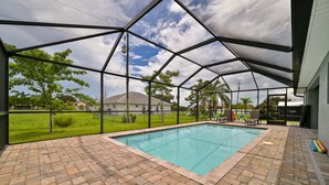 A covered outdoor swimming pool with a brick patio, surrounded by a metal fence, and a view of houses and greenery in the background. - Modern Coastal 3 Bedroom Villa with Salt Water Heated Pool & Office! (Cape Coral)