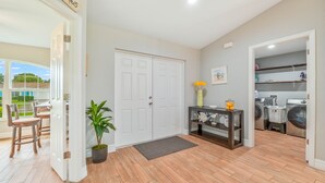 A spacious living area with a wooden floor, a white door, a potted plant, a console table with a vase, and a laundry area with washing machines. - Modern Coastal 3 Bedroom Villa with Salt Water Heated Pool & Office! (Cape Coral)