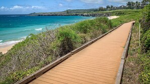 En träpromenad leder till en strand med kristallklart turkosfärgat vatten och en avlägsen utsikt över ett semesterområde.