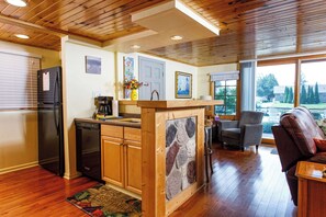 Una cocina con armarios de madera, un refrigerador, un lavavajillas, un protector contra salpicaduras de piedra y una zona de comedor con una silla y una mesa.