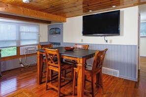 Ein Essbereich mit einem Holztisch und Stühlen, einem an der Wand befestigten Fernseher und einem Fenster mit Jalousien.
