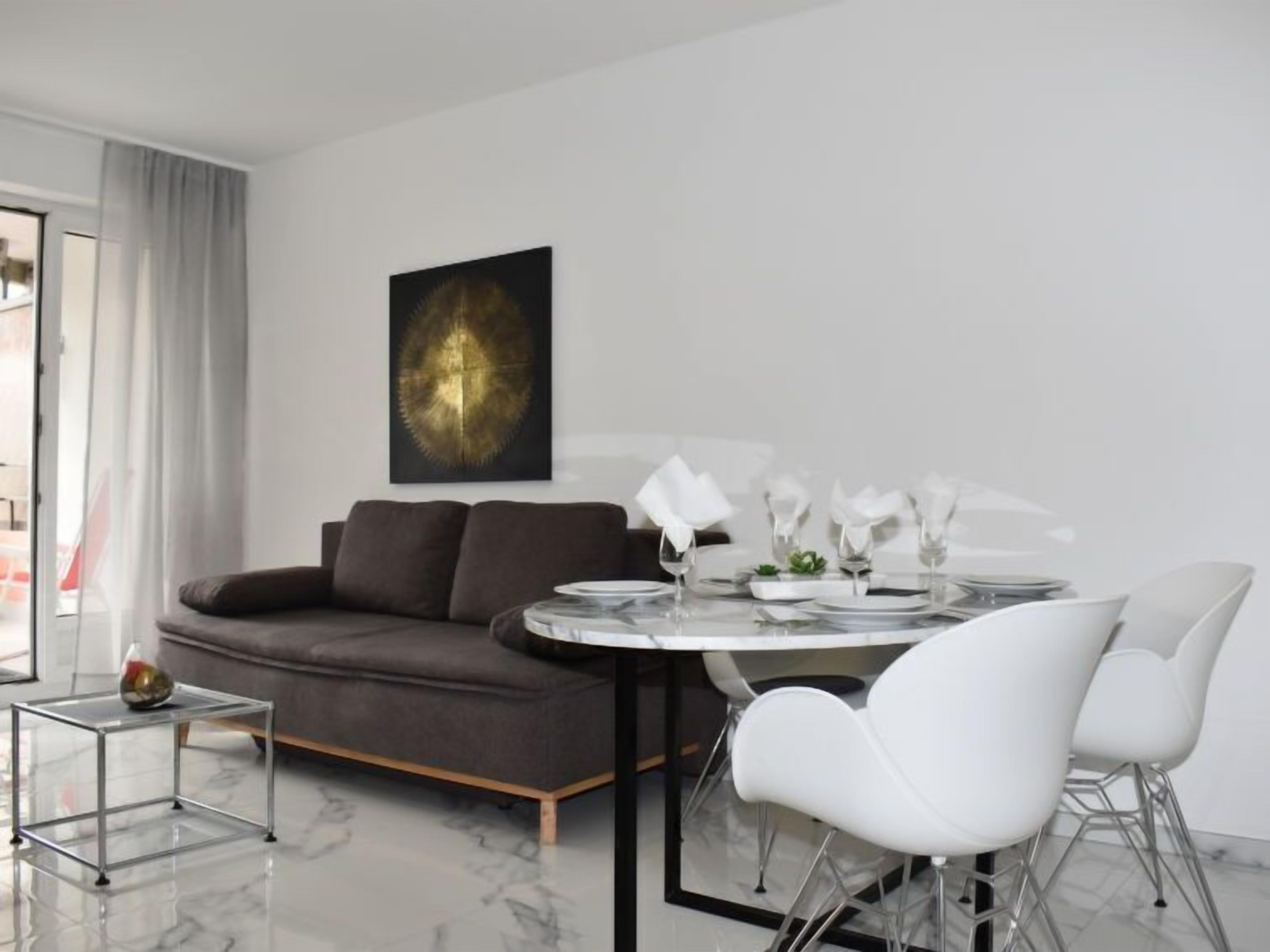 Una sala de estar moderna con un sofá gris, una mesa de comedor redonda con platos y vasos blancos, y un cuadro minimalista en la pared.