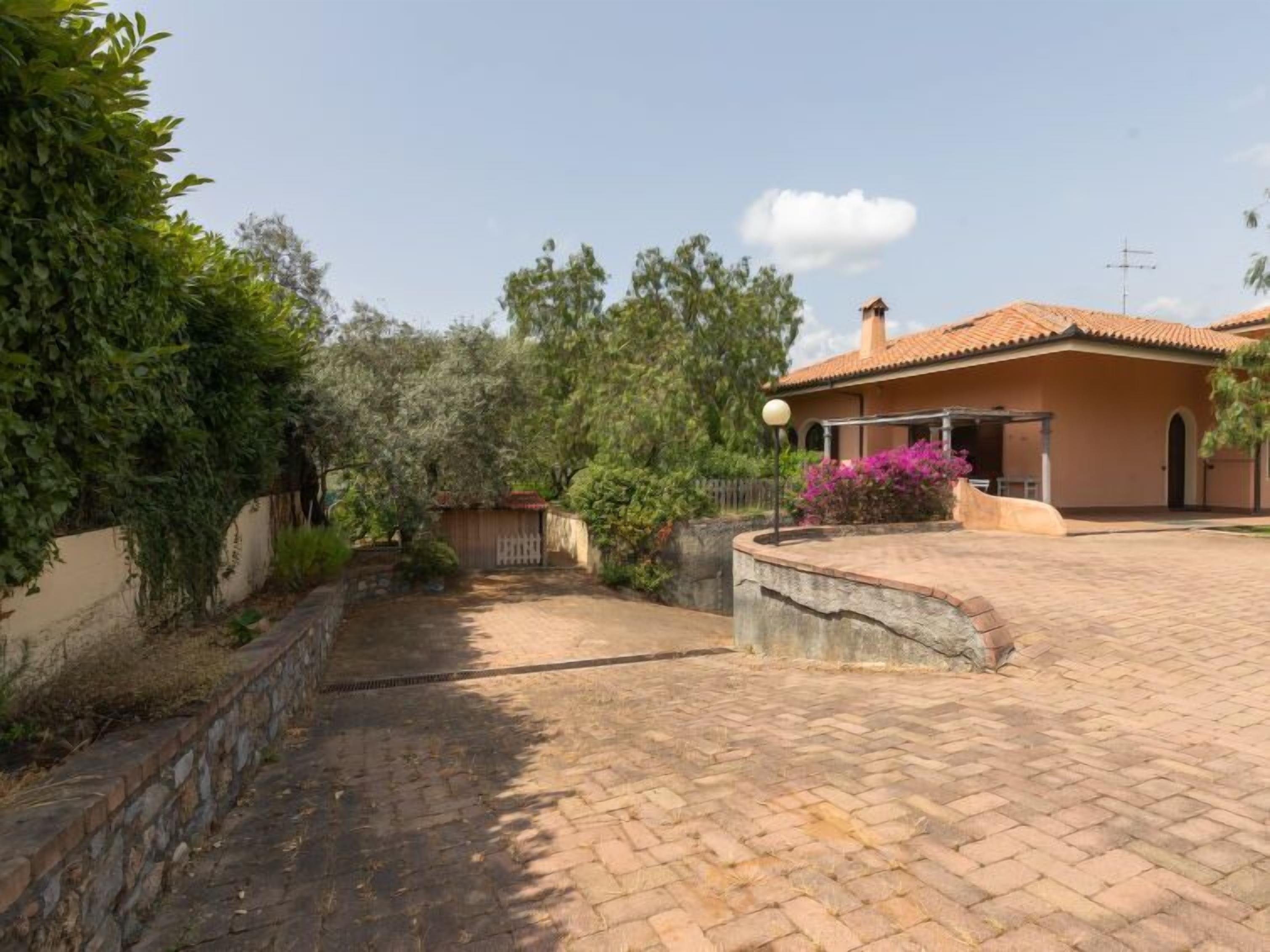 A house with a paved driveway, a stone wall, and a garden with trees and shrubs.
