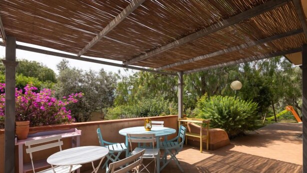 Een terras buiten met een overdekt gedeelte, een ronde tafel en opklapbare stoelen.