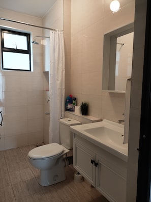 A bathroom with a toilet, sink, and shower. There is a window above the toilet and a mirror above the sink.