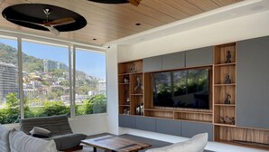 A modern living room with a large TV, grey sofas, wooden shelves, and a city view. - Great Comfort, Location, and Style Zona Romantica (AVIDA) (Puerto Vallarta)