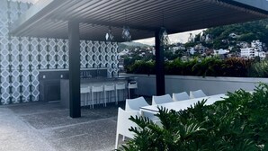 An outdoor patio with a modern design, featuring a pergola, seating area, and a view of a hillside town. - Great Comfort, Location, and Style Zona Romantica (AVIDA) (Puerto Vallarta)