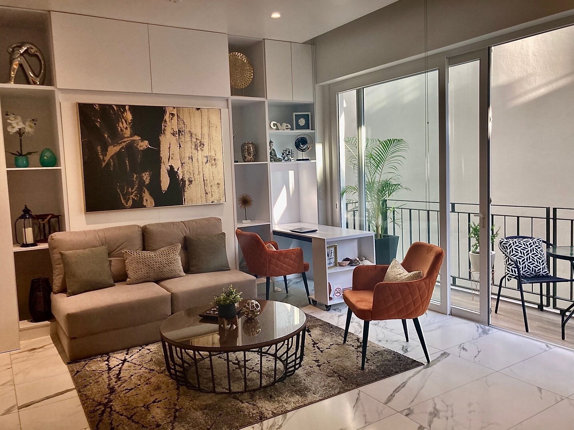 Una sala de estar moderna con un sofá, una mesa de centro y sillas, un cuadro grande y un balcón con plantas.