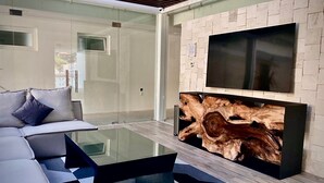 A modern living room with a glass coffee table, a sofa with cushions, a mounted flat-screen TV, and a wall with a wooden texture. - Great Comfort, Location, and Style Zona Romantica (AVIDA) (Puerto Vallarta)