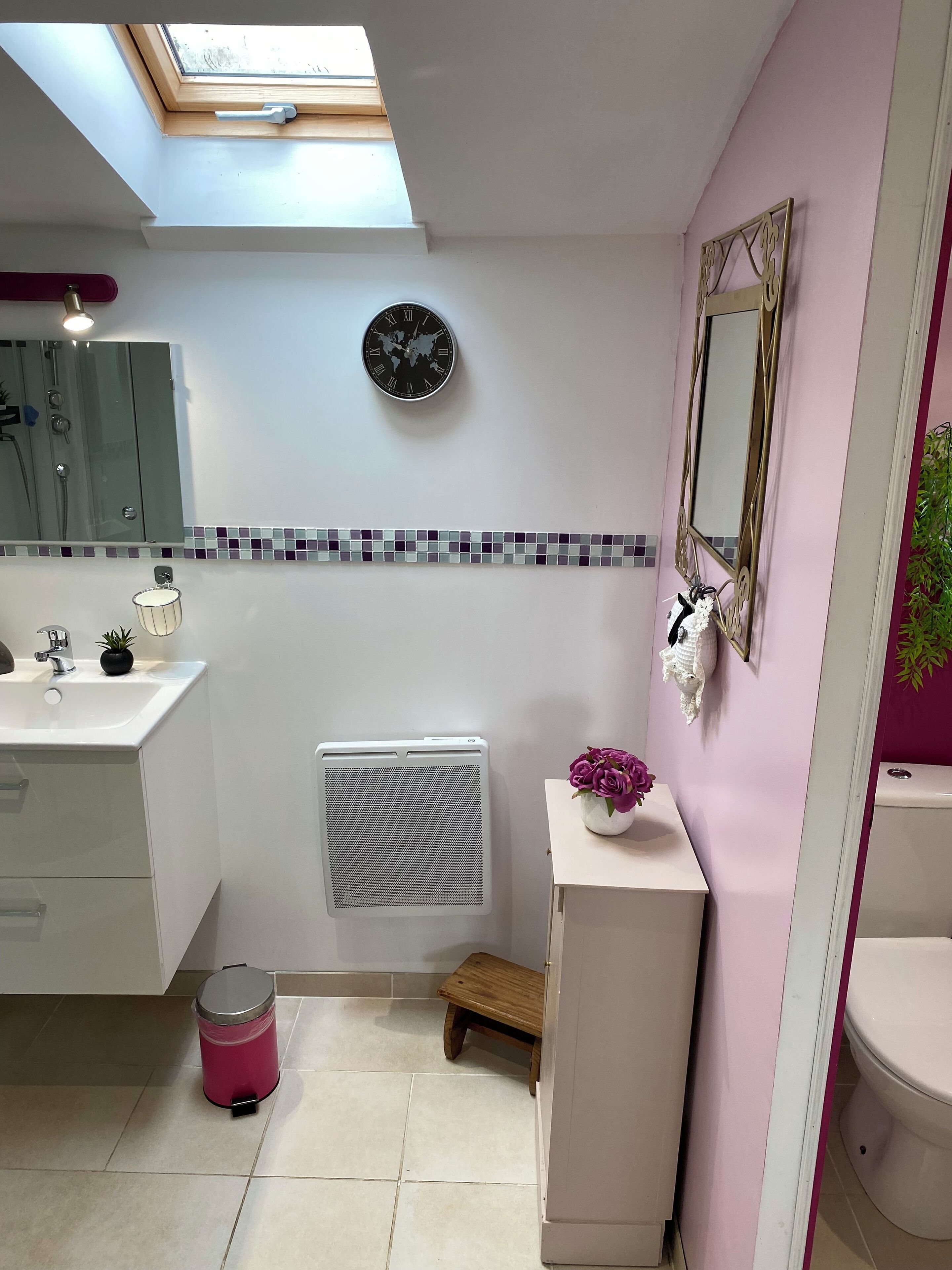 Une salle de bain avec un puits de lumière, une horloge indiquant l’heure dans différentes parties du monde, un radiateur, un petit tabouret et une poubelle.