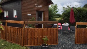 Una cabaña de madera con un cartel que dice "Le Chêne". Tiene una valla de madera, mobiliario exterior con mesas y sillas, y un paraguas rojo.